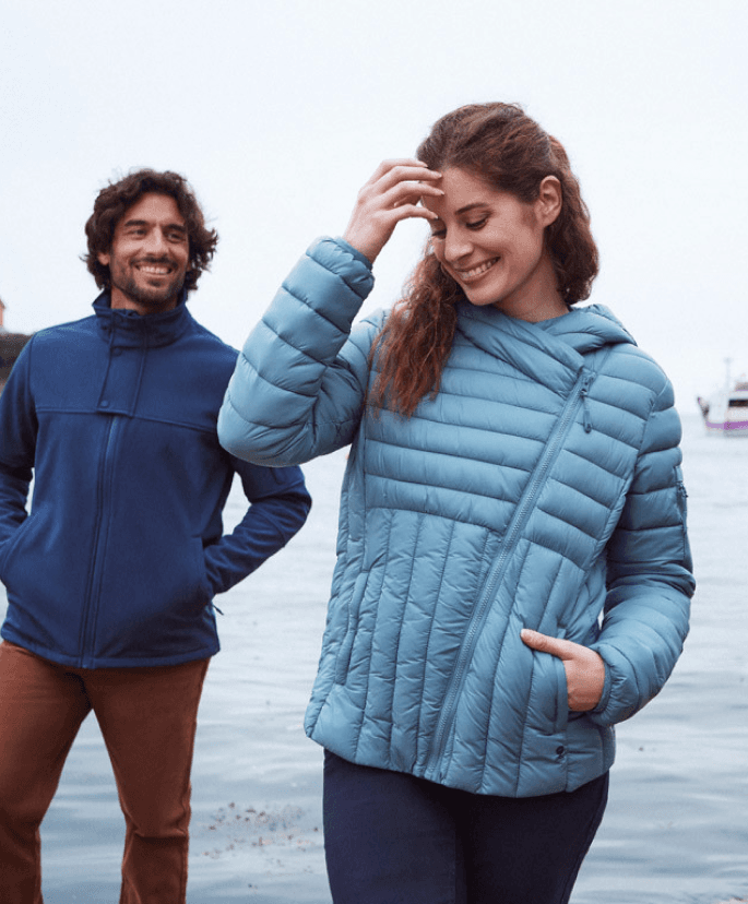 Une femme souriante portant une veste matelassée bleu clair se tient près d'un homme vêtu d'une veste bleue marine, dans un décor extérieur au bord de l'eau. Ils semblent profiter d'un moment détendu sous un ciel légèrement couvert.